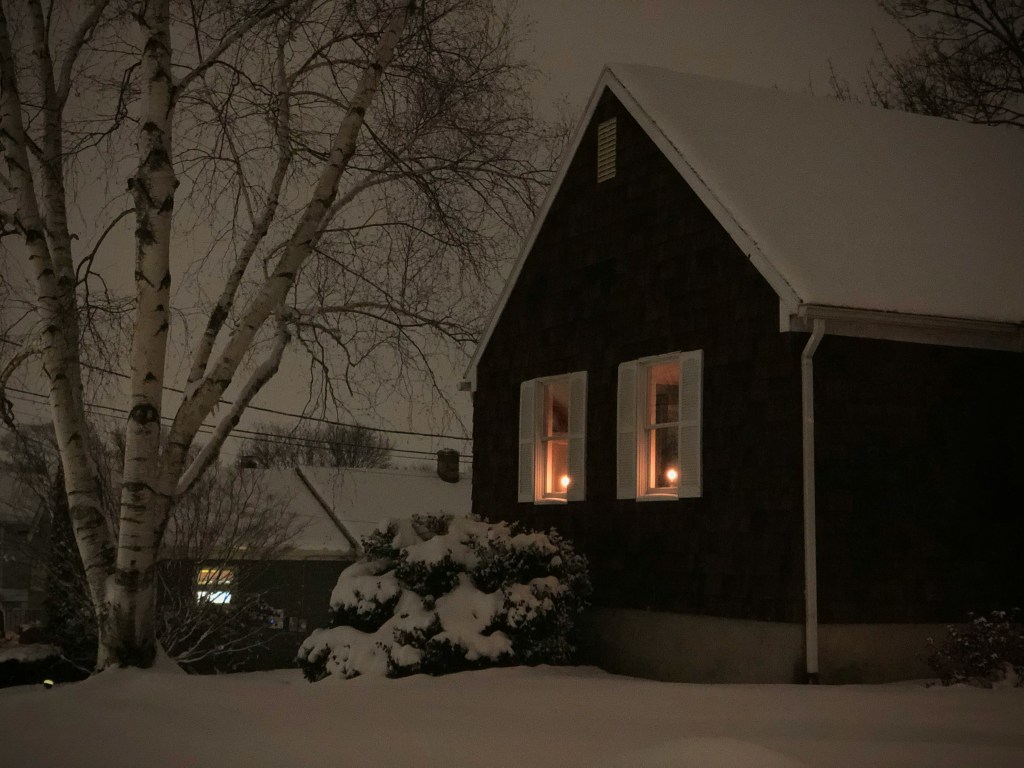 A low light image of a house in winter with snow around. There are  two windows, each with a candle illuminated in them.