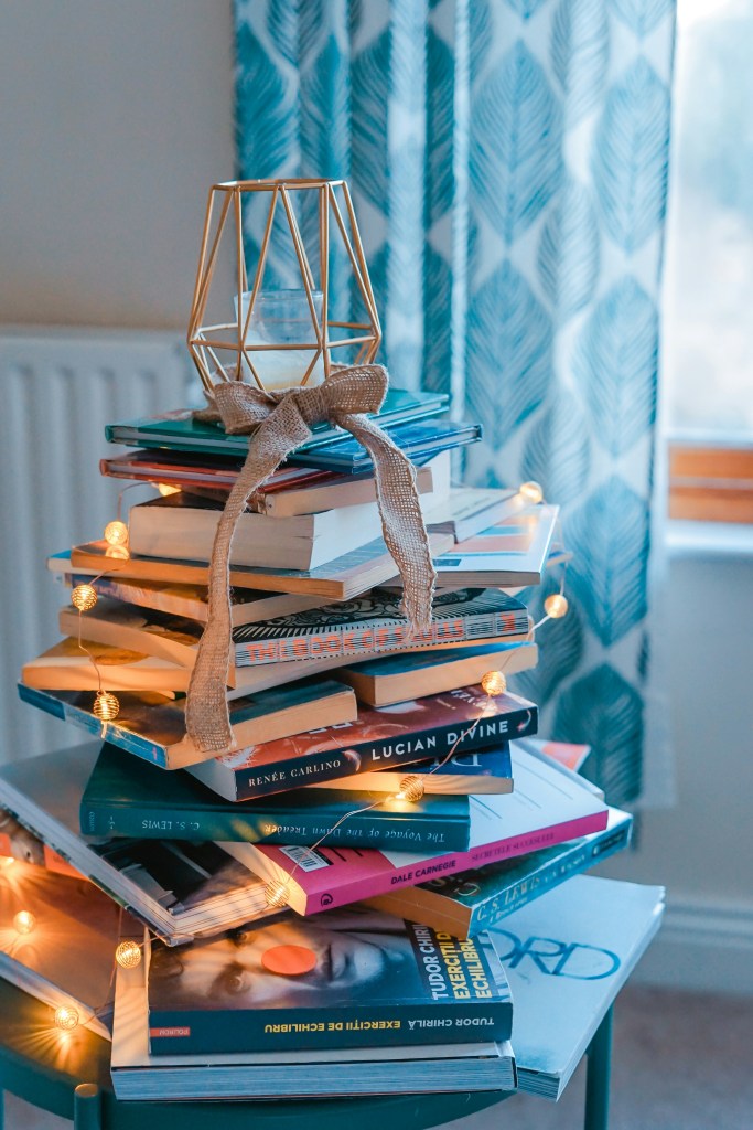 A stack of books with a string of small white lights and a candle in a geometric holder on top.